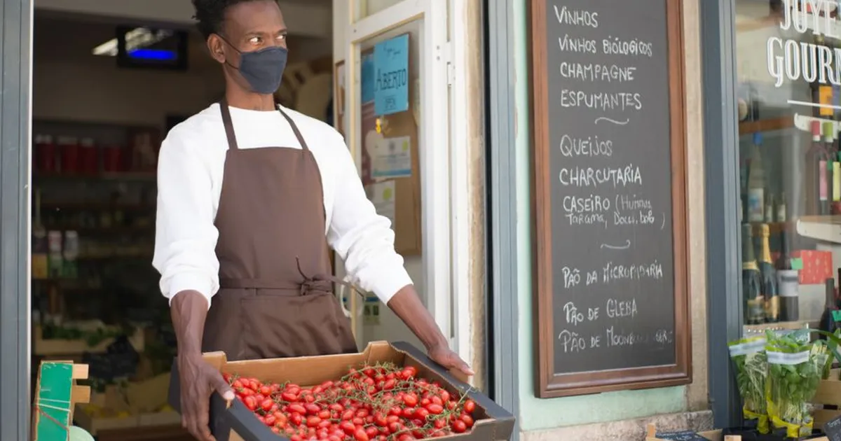 A vendor wearing a mask holds a crate of fresh tomatoes outside a Portuguese market.