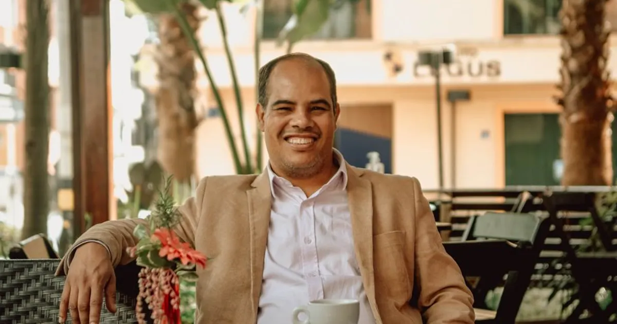 Balding man in a suit jacket smiling while enjoying a coffee in an outdoor café setting.
