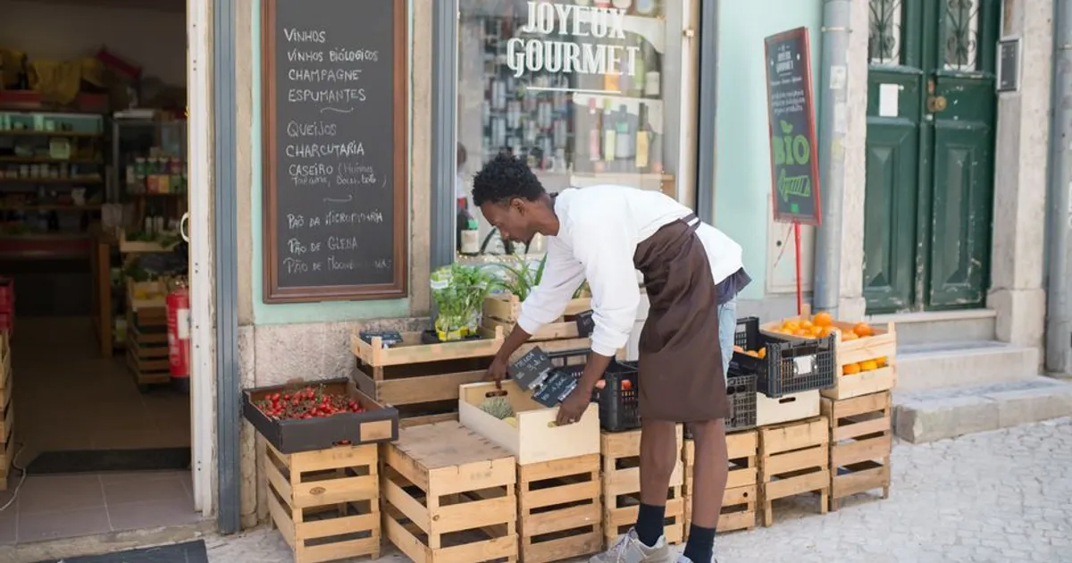 A worker arranges fruit crates outside a grocery store in Portugal, adding vibrant oranges and strawberries.