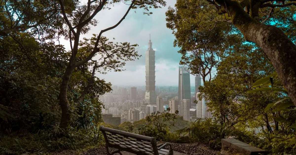 A picturesque view of Taipei 101 skyscraper framed by lush greenery and a park bench.