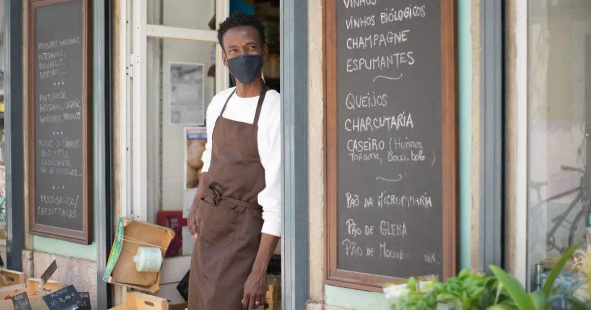 Portrait of a masked shopkeeper standing in a Portuguese store doorway with chalkboard menus.