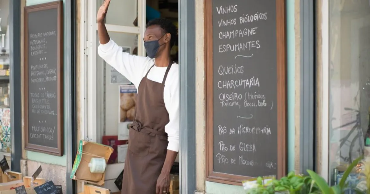 A grocery store clerk wearing an apron and face mask warmly waves from the storefront during pandemic times.