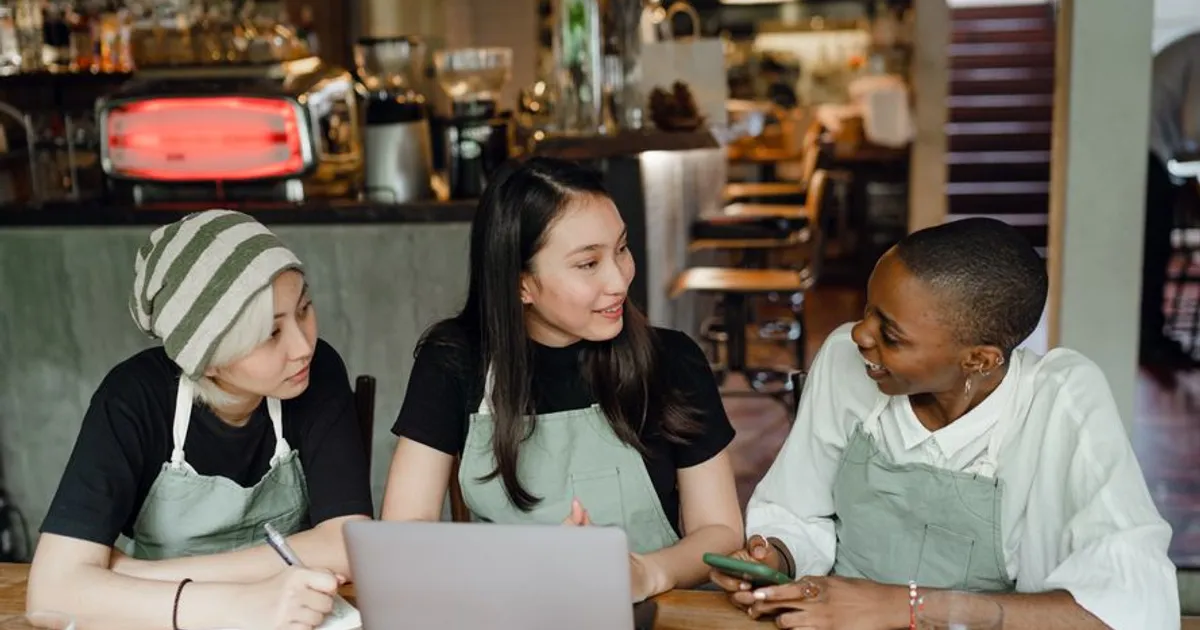 Positive young multiethnic waitresses in aprons sitting at wooden table in modern cafe and working on startup project on contemporary netbook