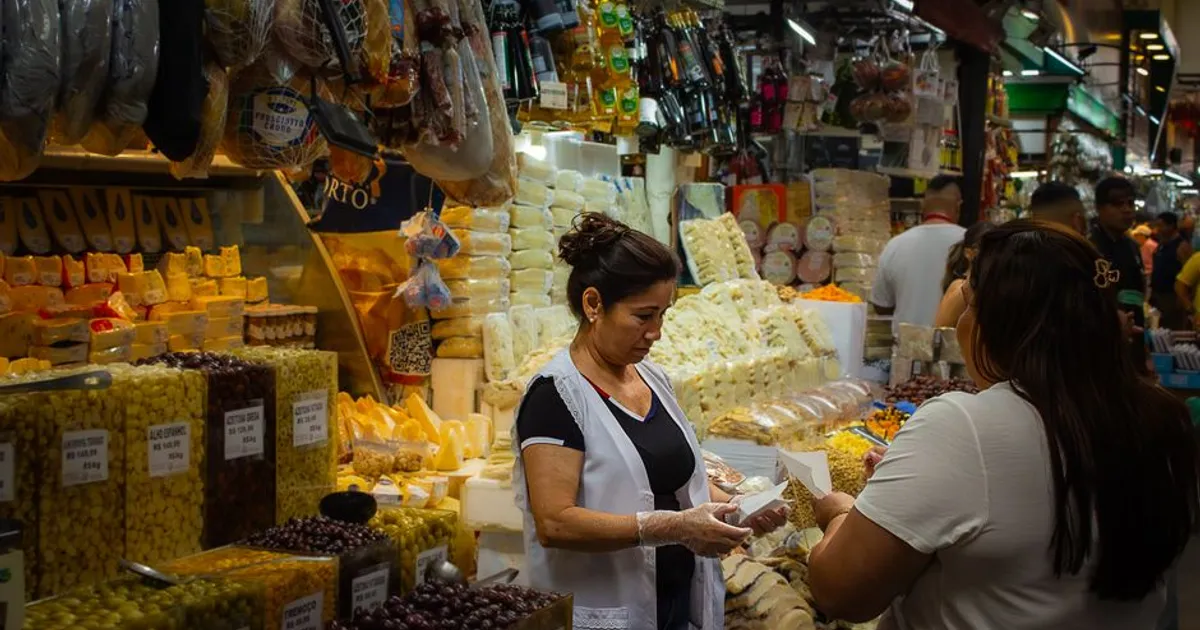 A crowded indoor market with a cheese seller attending customers amidst a variety of gourmet foods.